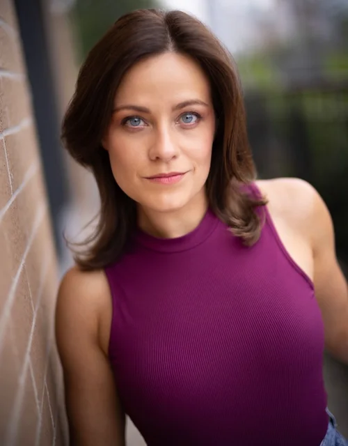 Person with short brown hair, blue eyes, purple sleeveless top posing next to a brick wall