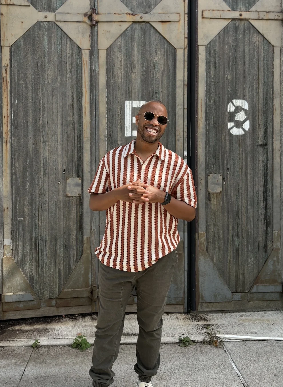 Marcel Stewart standing in front of a large door wearing a striped red and white shirt and green pants and sunglasses